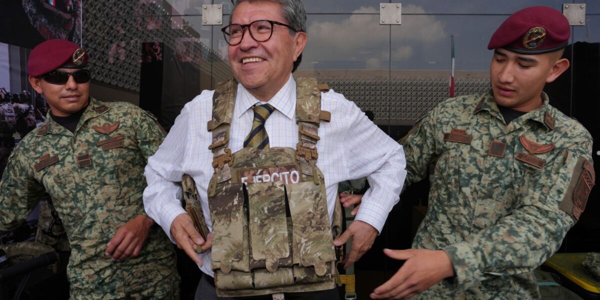 Ricardo Monreal prueba un chaleco táctico acompañado de dos militares durante una exhibición.